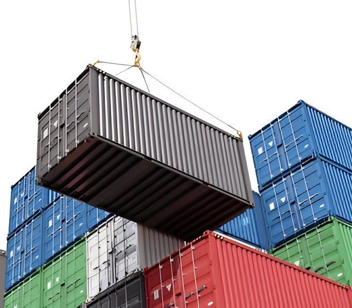Gray shipping container being lifted by a crane above stacks of colorful containers