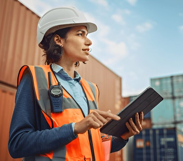 Worker in a hard hat and hi-vis vest using a tablet in a shipping container yard