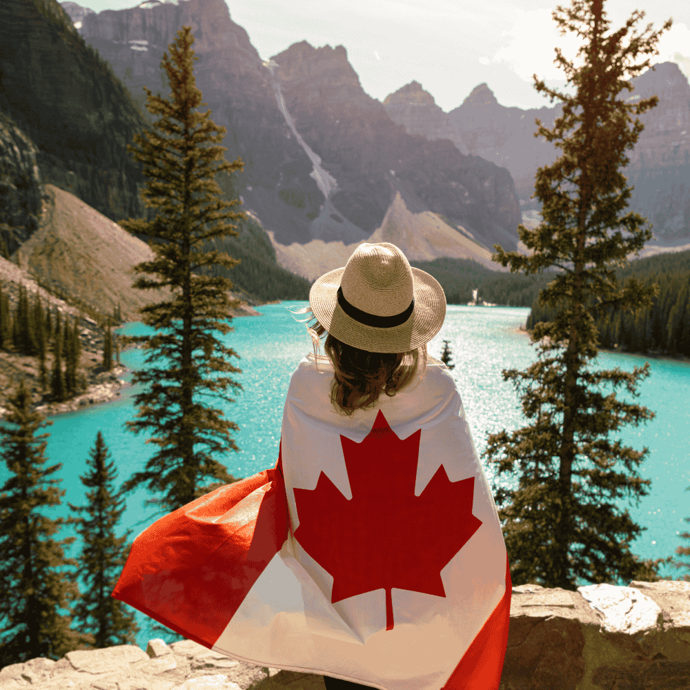 Person wrapped in a Canadian flag looking over a turquoise lake and Rocky Mountains (Banff/Moraine Lake scene)