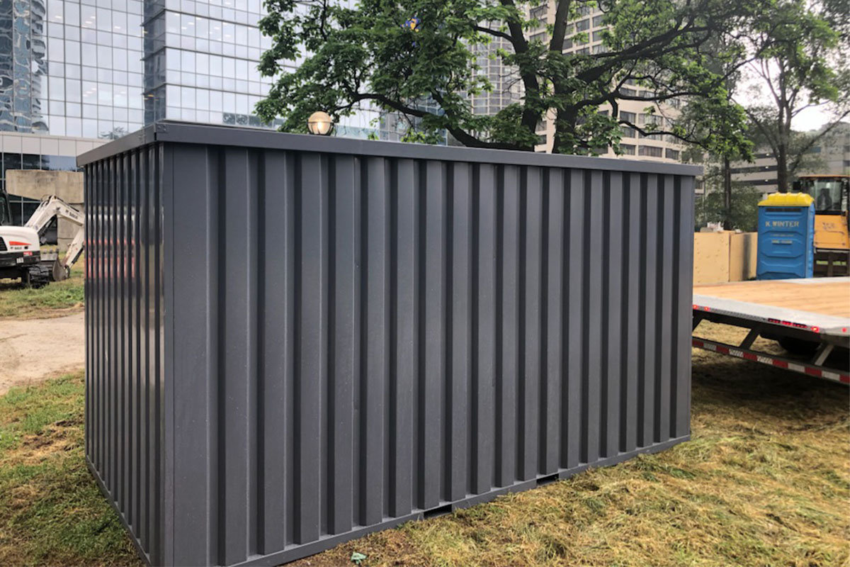 Dark grey steel storage container placed on a residential lawn beside a flatbed delivery truck in an urban neighborhood
