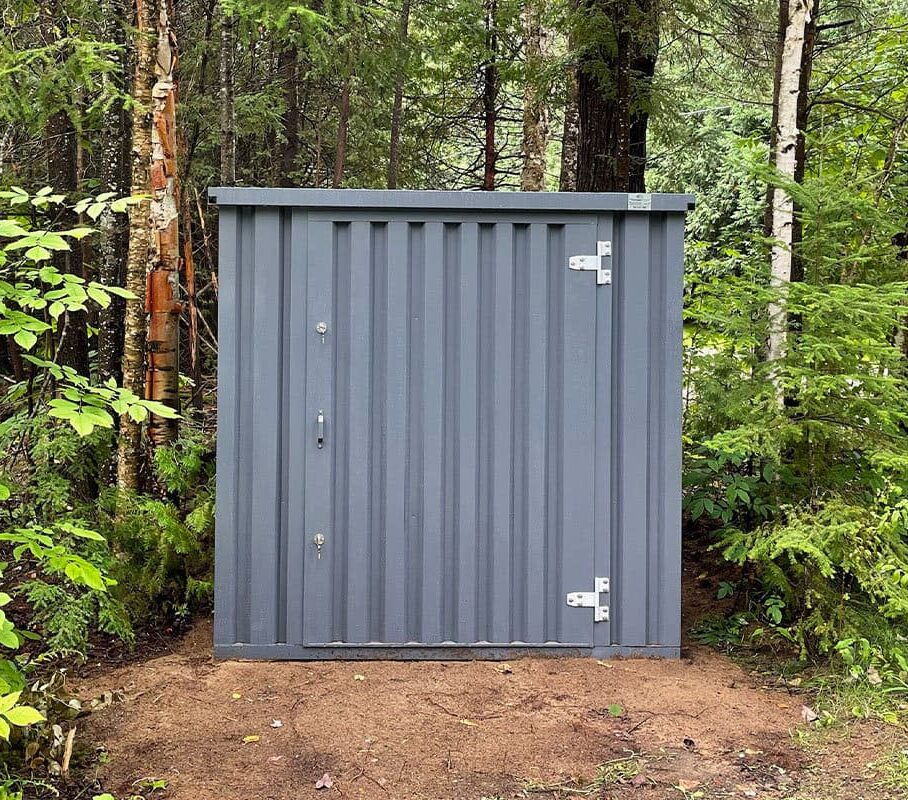 Grey flat-pack steel storage shed in a forest clearing, front view with locking door