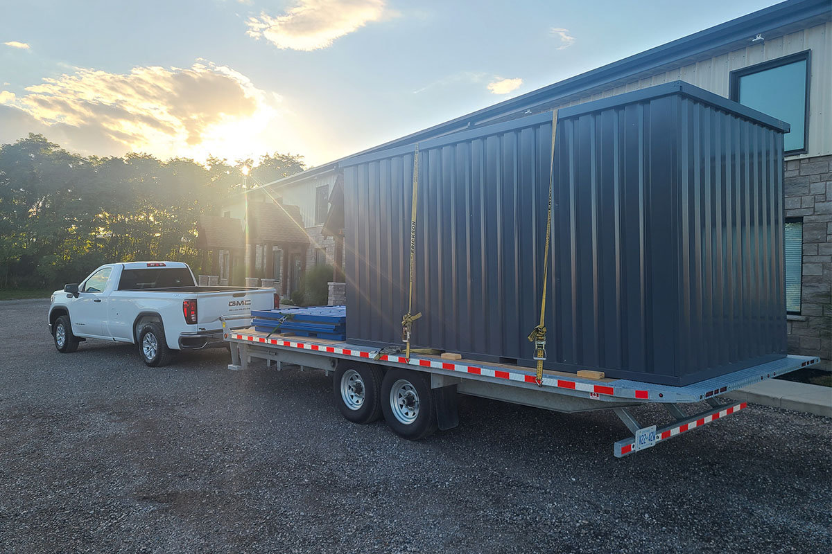 Dark-grey backyard storage container strapped on a flatbed trailer behind a white pickup truck at sunset outside a home