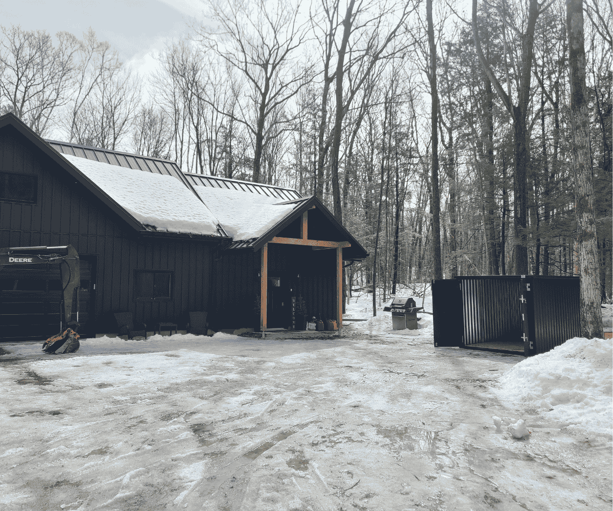 Open dark gray storage container beside a modern cottage in snowy woods — winter placement by Alloy Storage