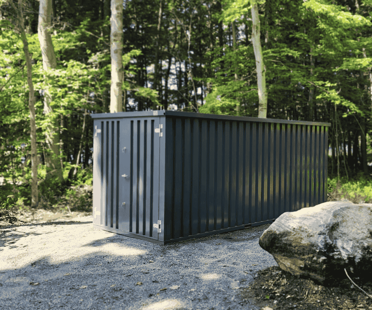 Dark gray steel storage container on a gravel pad in the woods near a cottage, secure seasonal gear storage — Alloy Storage